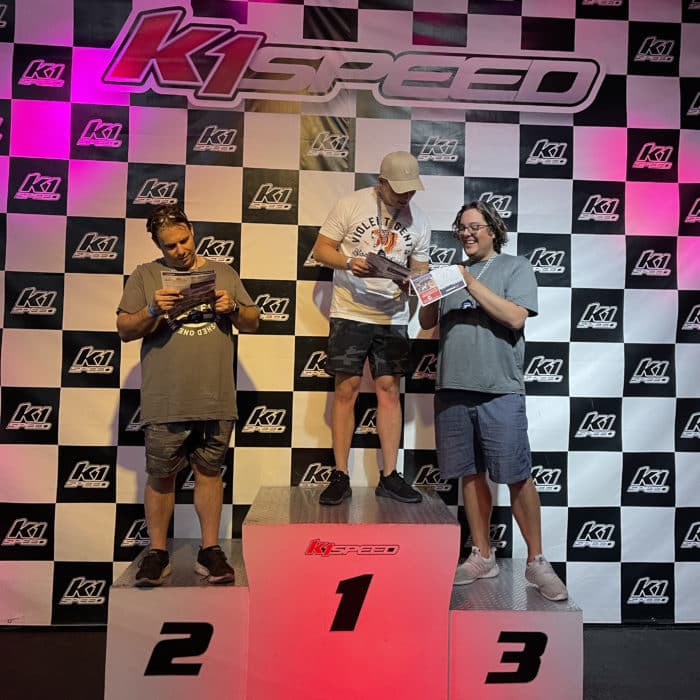 Three Kingsmen employees standing on the first, second, and third placement blocks holding their medals at K1 Indoor Racing