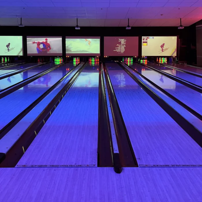 Several bowling lanes with large TV screens above each lane and a deep purple light glowing over the alleys